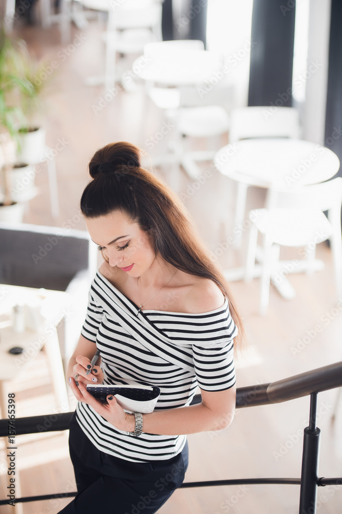 Attractive young woman writing in her notebook while standing on stairs in a cafe and looking down.