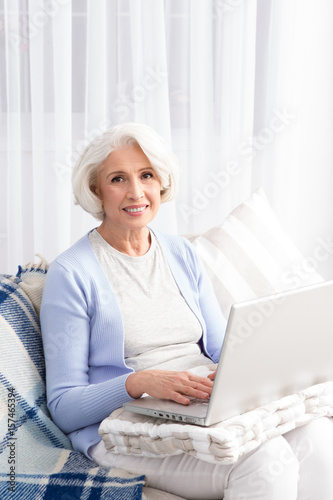 Elderly woman keeping record of finances in laptop. Happy woman smiling for camera while sitting on sofa or couch. Computer technologies concept.