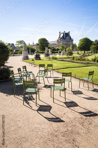 View of the Tuileries garden in Paris by a sunny morning with the famous metal lawn chairs in the foreground and the Flore pavilion of the Louvre palace in the background © olrat