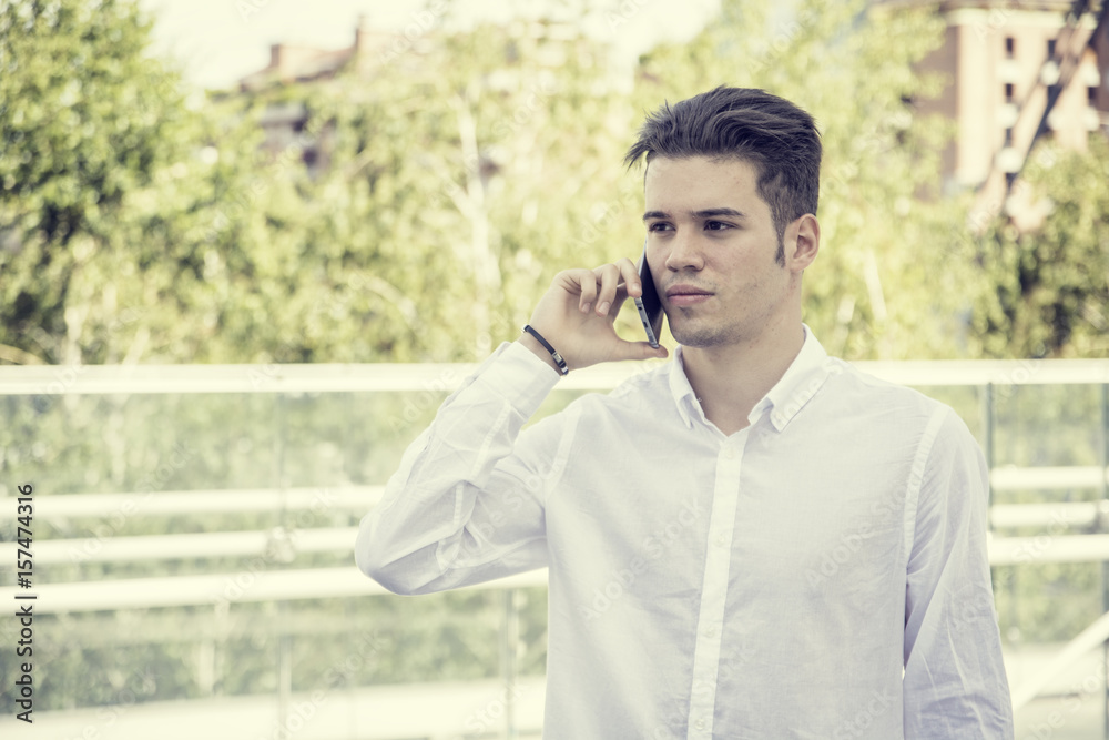 Handsome trendy young man wearing white shirt standing and talking on cell phone, outdoor in city setting in day shot Handsome trendy young man wearing white shirt standing and talking on cell phone, outdoor in city setting in day shot