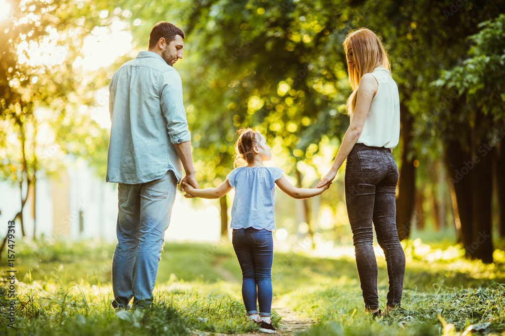 Obraz premium Happy family in the park. Mom, dad and baby happy walk at sunset. The concept of a happy family. Parents hold the baby's hands.
