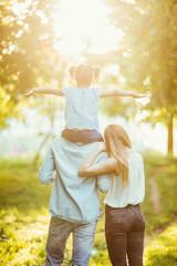 Happy family of three persons walking the grass in the park on sunset. happy daughter flying on father back