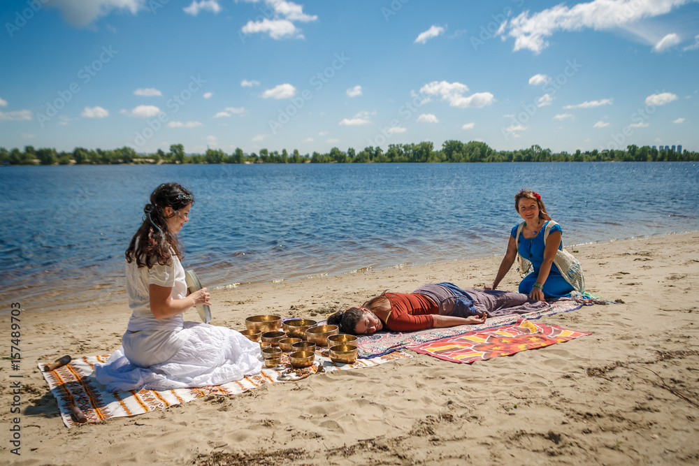 Beautiful female receiving energy sound massage with singing bowls and body massage on a river bank at spring sunny day