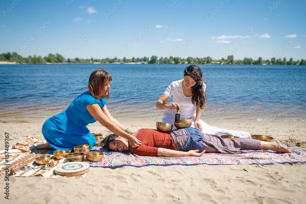 Obraz premium Beautiful female receiving energy sound massage with singing bowls and body massage on a river bank at spring sunny day