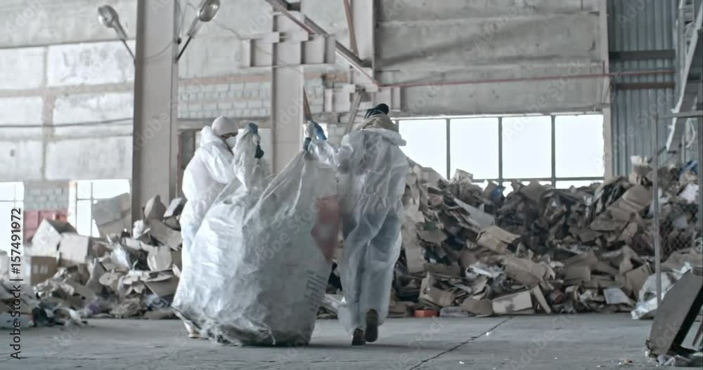 Slow motion dolly shot of garbage recycling factory workers in protective coveralls and masks carrying bag of litter towards cardboard waste as their colleague sorting plastic litter 