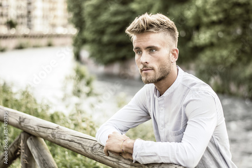Blondish, blue eyed young man by river at sunset, thinking, looking away