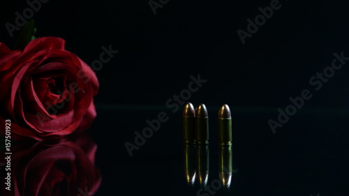 Bullets and a red rose on the black mirror floor. Bullets are a projectile expelled from the barrel of a firearm.