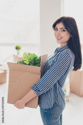 Successful young woman is moving to new nice place and holding box with her belongings. The room is very light and bright, she is wearing cassual outfit