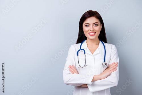 Portrait of cute doctor in white coat standing with crossed hands against gray background