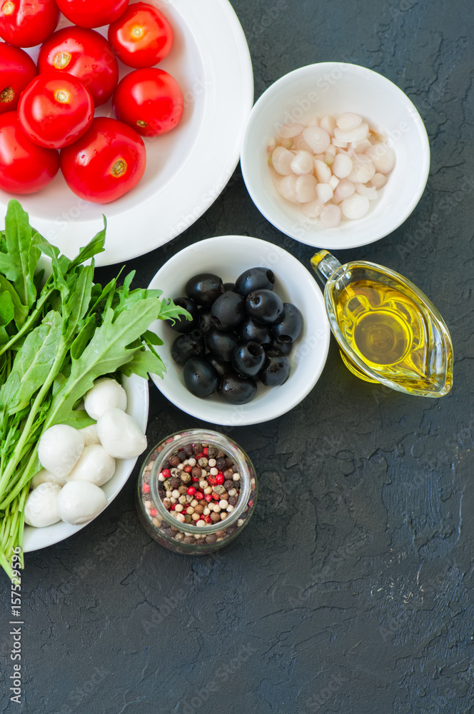 Ingredients for healthy salad. Tomatoes, rucola, spring onion, mozzarella, olives, garlic and pepper with olive oil on a black slate background.
