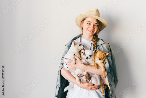 Young smiling happy hipster girl in straw hat, jeans jacket with russian style tress and upsweep holding in hands four chihuahua puppies dogs with funny emorional faces. Coiffure. Looking at camera. photo