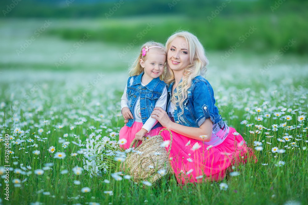 Fototapeta premium Mom and daughter on a picnic in the chamomile field. Two beautiful blondes wearing jeans jacket and pink skirt Mother and baby girl embracing in the chamomile field with basket of bouquet dandalion