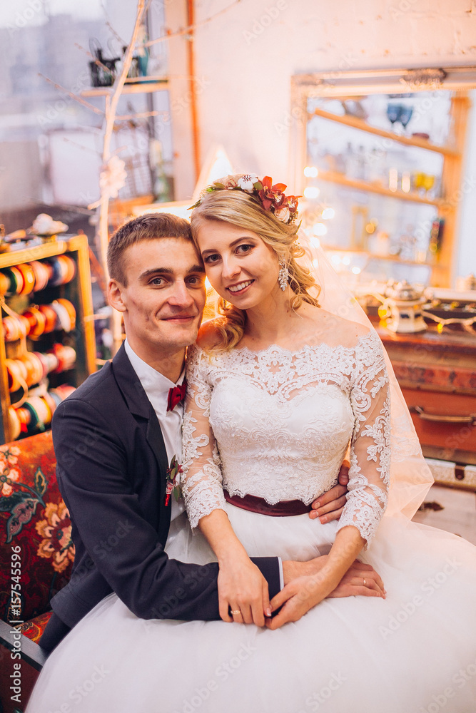 Naklejka premium Bride in beautiful dress and groom in black suit sitting on sofa indoors in white studio interior like at home. Trendy wedding style. Groom.bride hugging