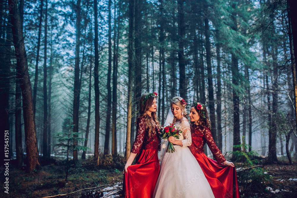 Bride and bridesmaids in red dresses stand on the path in pine forest.