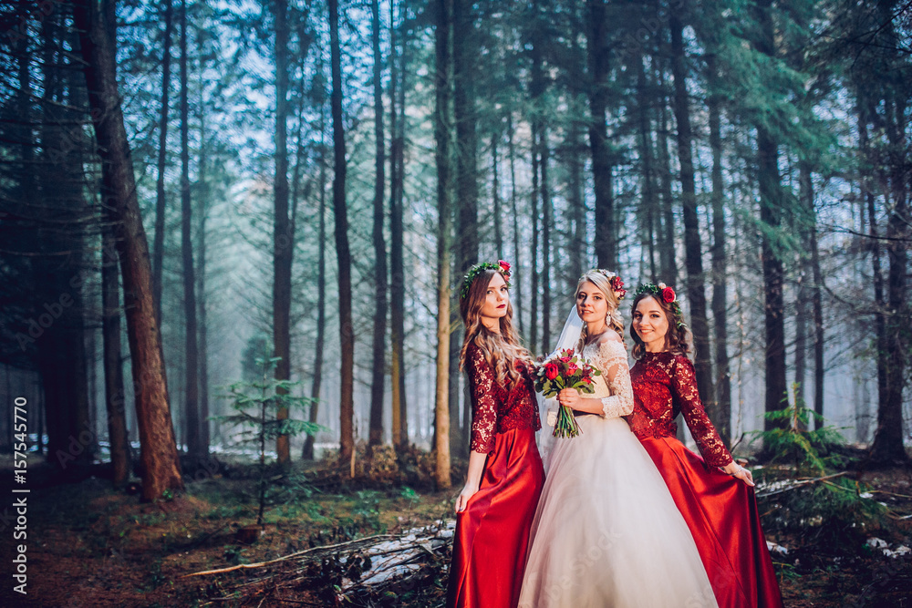 Bride and bridesmaids in red dresses stand on the path in pine forest.