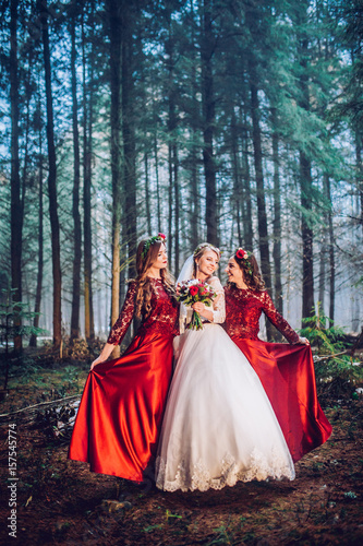 Bride and bridesmaids in red dresses stand on the path in pine forest.