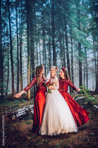 Bride and bridesmaids in red dresses stand on the path in pine forest.