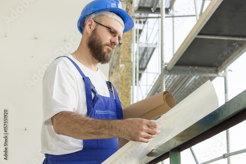 Pracownik budowlany w stroju roboczym i w kasku stoi na dużej wysokości na budowie i przegląda plany techniczne.
