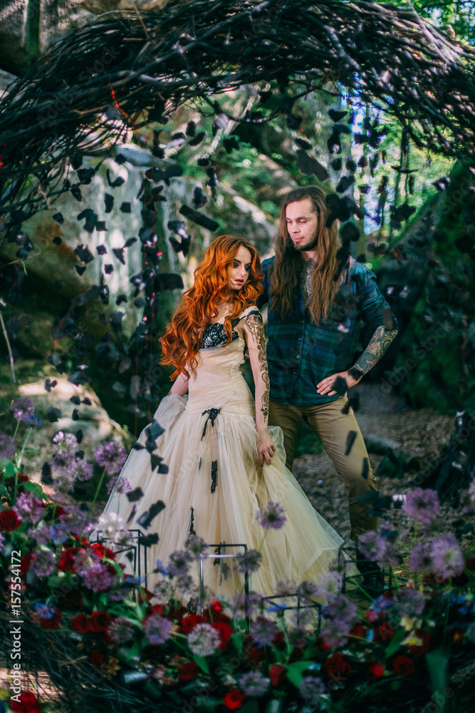 Fototapeta premium Beautiful young couple together near dark circle arch with flowers. Man with tattoo and long hair with beard. Woman with red hair. Fairytale concept.