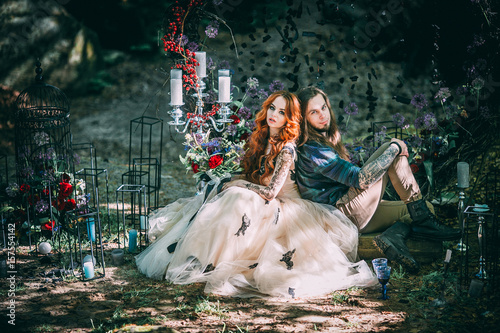 Obraz na płótnie Beautiful young couple together near dark circle arch with flowers. Man with tattoo and long hair with beard. Woman with red hair. Fairytale concept. w salonie