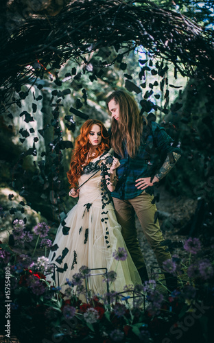 Beautiful young couple together near dark circle arch with flowers. Man with tattoo and long hair with beard. Woman with red hair. Fairytale concept.