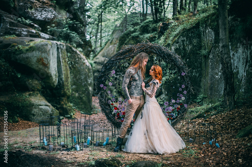 Obraz na płótnie Beautiful young couple together near dark circle arch with flowers. Man with tattoo and long hair with beard. Woman with red hair. Fairytale concept. w salonie