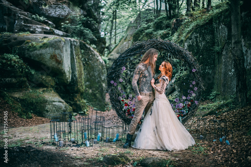 Beautiful young couple together near dark circle arch with flowers. Man with tattoo and long hair with beard. Woman with red hair. Fairytale concept.