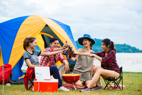 Group of man and woman enjoy camping picnic and barbecue at lake with tents in background. Young mixed race Asian woman and man. Young people's hands toasting and cheering bottles of beer. © tirachard