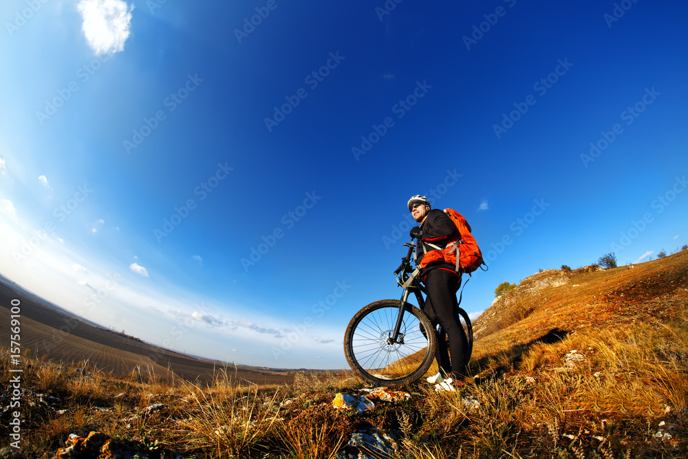 Naklejka premium Low, wide angle portrait against blue sky of mountain biker going downhill. Cyclist in black sport equipment and helmet