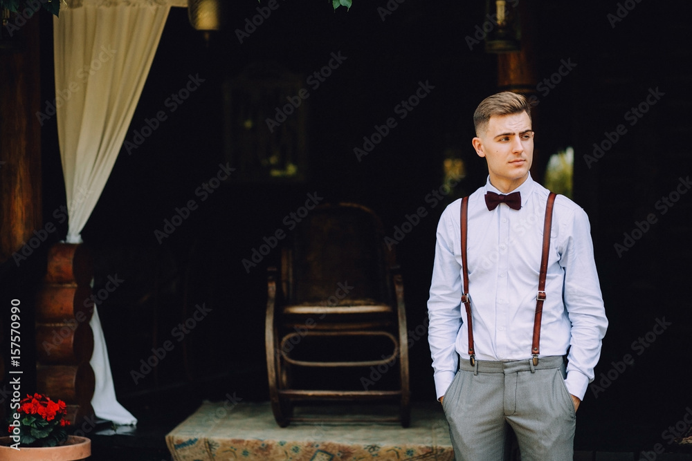 Ταπετσαρία A handsome young man in a shirt, a bow tie, trousers and suspenders stands on the veranda of the house
