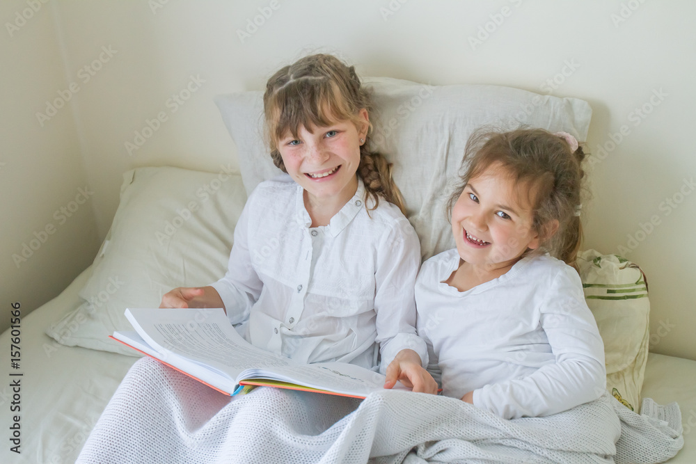Obraz premium indoor portrait of young european girls - two sisters - lying in bed and reading a book