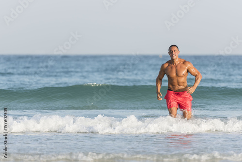 Obraz na płótnie Mature older muscular fit Arab Male running out of the ocean on the beach, shirtless, wearing red shorts w salonie