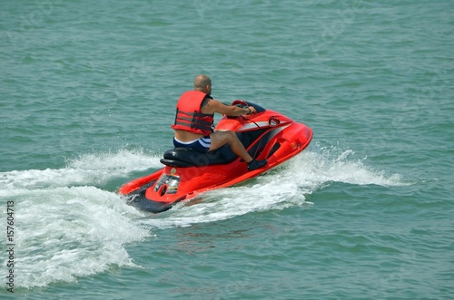 A baldheaded man wearing a red life jacket and riding a red jet ski speeding across the florida intra-coastal waterway off Miami Beach.