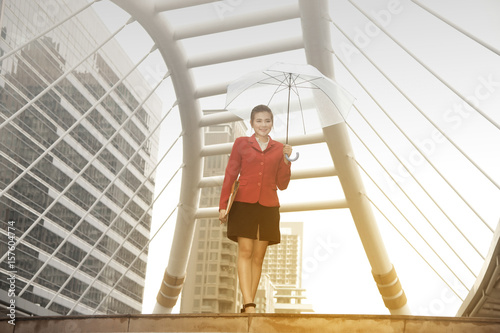 Thai pretty woman in red office suit holding transparent umbrella walking on bridge crossing landmark in Bangkok,Thailand.