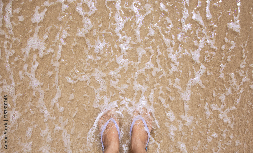Fototapeta premium Top view of woman feet wearing a blue slipper on ocean sandy beach, relaxing concept