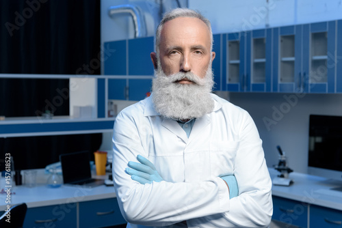 serious grey haired scientist in lab coat looking at camera with arms crossed