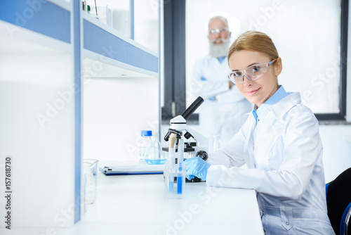 Smiling middle aged scientist in protective eyewear working with microscope and colleague standing behind