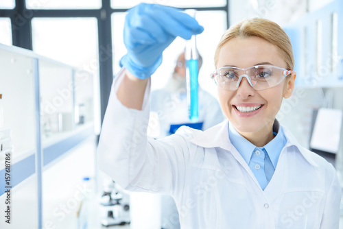 Smiling chemist in protective eyeglasses holding test tube with reagent in lab