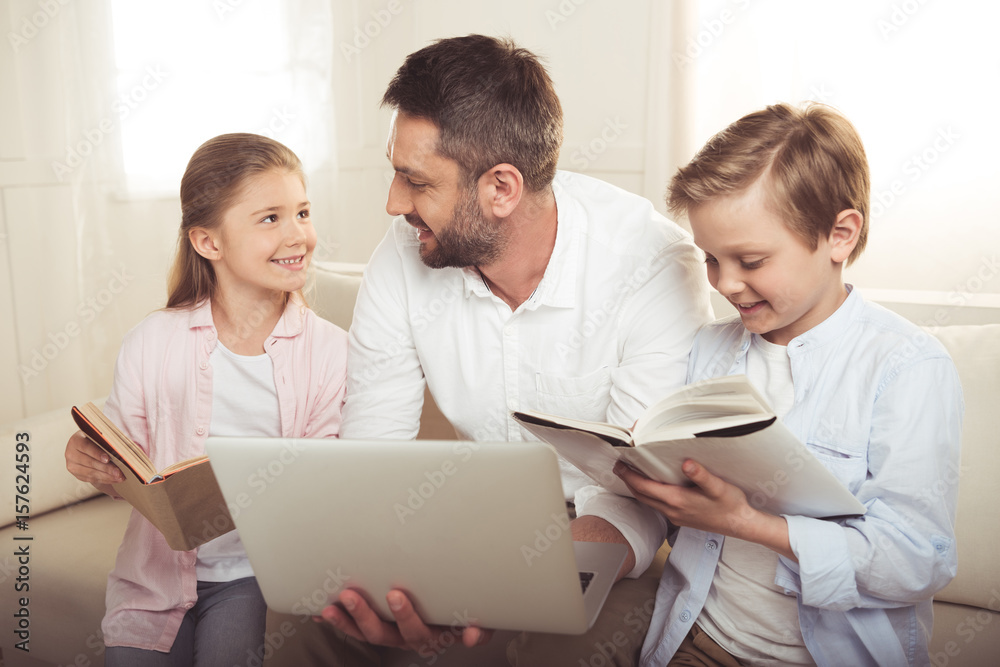 cheerful father using laptop while his daughter and son reading books and sitting on sofa cheerful father using laptop while his daughter and son reading books and sitting on sofa