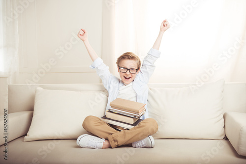Happy little boy with pile of books sitting on sofa with raised hands