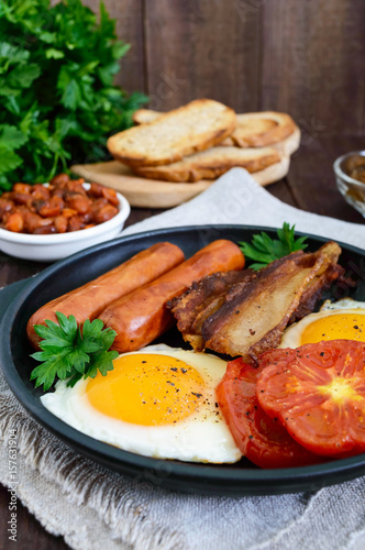 English breakfast: sausages, bacon, tomatoes, egg, beans in sauce, fried mushrooms, toast on a dark wooden background. Vertical view. Traditional classic food of England.