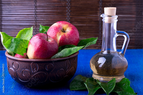 Apple cider vinegar in glass bottle on blue background. Red apples in brown bowl.