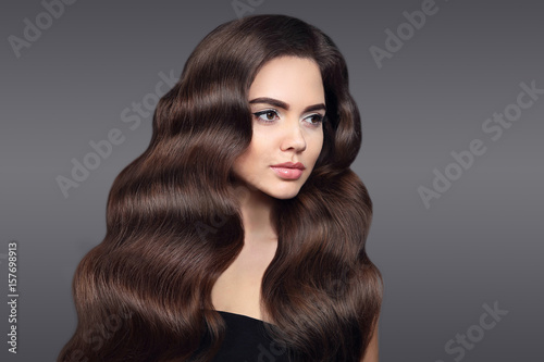 Healthy hair. Beautiful Brunette girl portrait with long shiny wavy hair. Attractive model with curly hairstyle and fresh makeup isolated on studio dark background. Shampoo care product.