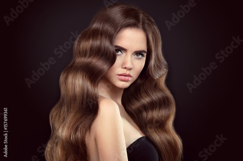 Healthy hair. Brunette girl portrait with long shiny wavy hair. Beautiful model with curly hairstyle and eyeliner makeup isolated on studio black background.