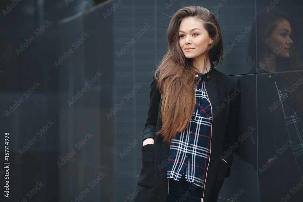 A young leader,a beautiful modern woman,a brunette with long straight hair,is dressed in a black stylish jacket and plaid shirt,posing standing outdoors near the mirror wall of the office A young leader,a beautiful modern woman,a brunette with long straight hair,is dressed in a black stylish jacket and plaid shirt,posing standing outdoors near the mirror wall of the office