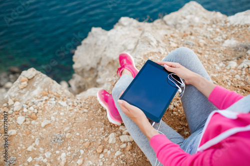 Woman in pink sneakers,a sports jacket pink,sitting in the open air, on the high rocky shore, against the blue of the ocean,ready to listen to music on your tablet through the white headphones