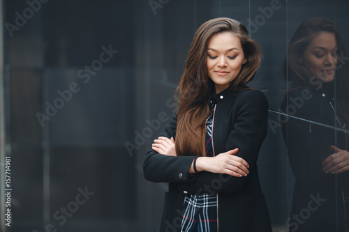 A young leader,a beautiful modern woman,a brunette with long straight hair,is dressed in a black stylish jacket and plaid shirt,posing standing outdoors near the mirror wall of the office