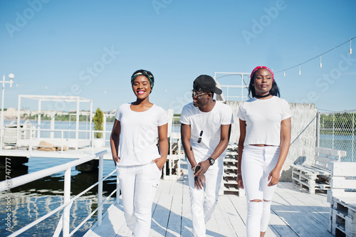 Three stylish african american friends, wear on white clothes at pier on beach. Street fashion of young black people. Black man with two african girls. © AS Photo Family