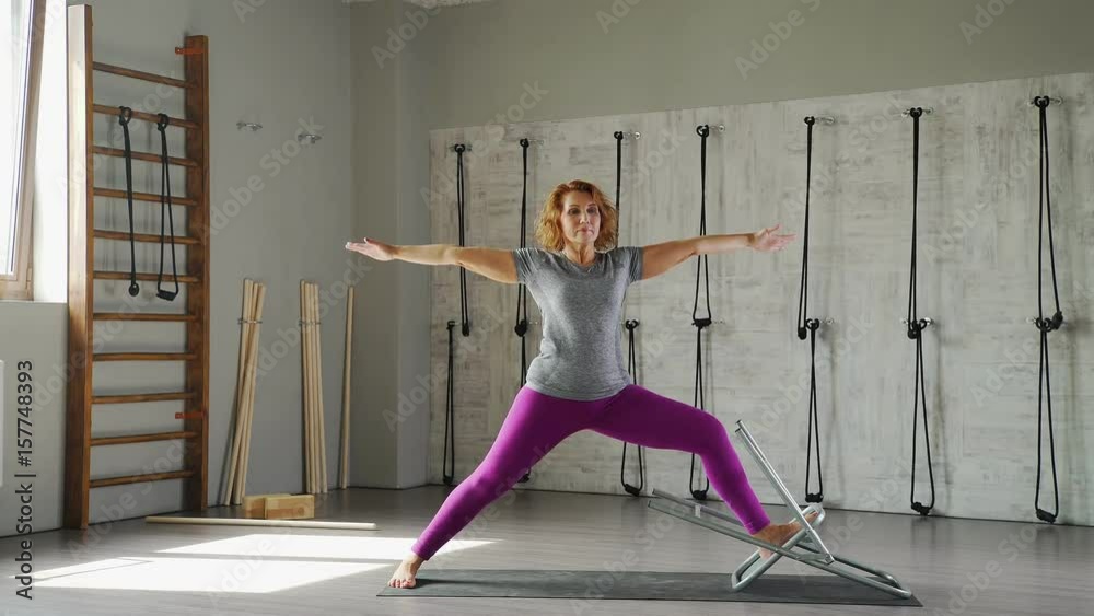 A mature woman practices yoga and performs asana the pose of a warrior . An adult female yogi makes an asana using a chair. A bright woman 40-45 years old is engaged in yoga in a bright studio. A mature woman practices yoga and performs asana the pose of a warrior . An adult female yogi makes an asana using a chair. A bright woman 40-45 years old is engaged in yoga in a bright studio.
