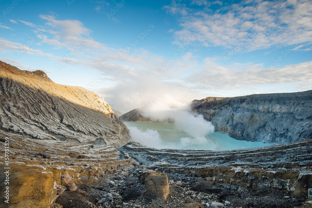 Obraz premium Ijen volcano complex in the morning. Sun shines on the mountain rim steam and smoke flying up the blue sky. Crater lake with turquoise water down.